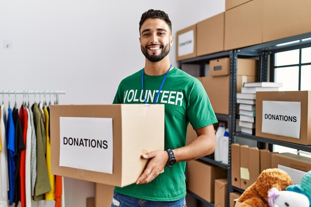Giovane uomo arabo che indossa un'uniforme da volontario che tiene una scatola per le donazioni al centro di beneficenza