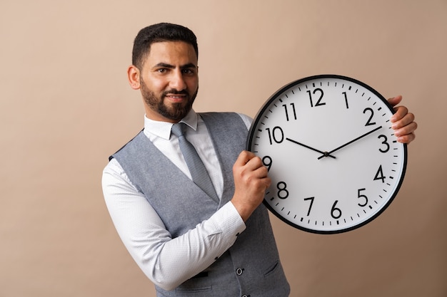 Giovane uomo arabo che tiene in mano un grande orologio da parete su sfondo beige