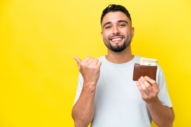 Giovane uomo arabo che tiene un portafoglio isolato su sfondo giallo che punta verso il lato per presentare un prodotto