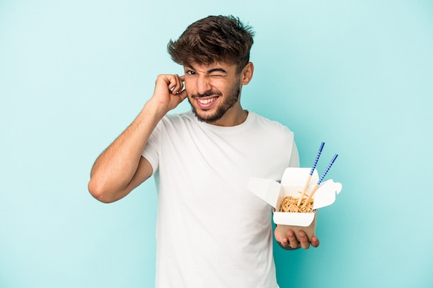 Giovane uomo arabo che tiene una pasta da asporto isolata su sfondo blu che copre le orecchie con le mani.