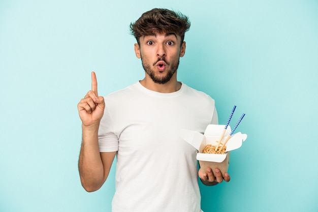 Giovane uomo arabo che tiene una pasta da asporto isolata su sfondo blu con qualche grande idea, concetto di creatività.