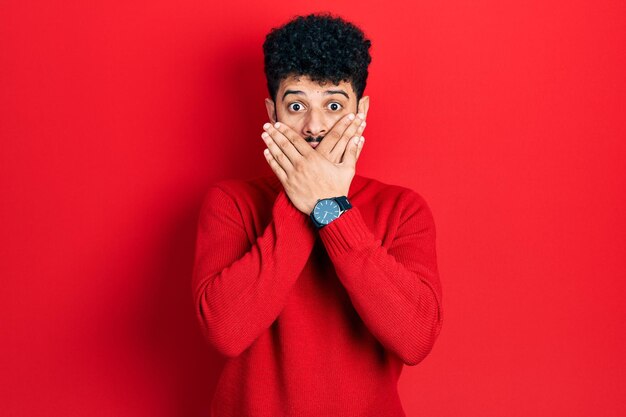 Giovane uomo arabo con la barba che indossa un maglione rosso casual scioccato coprendo la bocca con le mani per errore. concetto segreto.