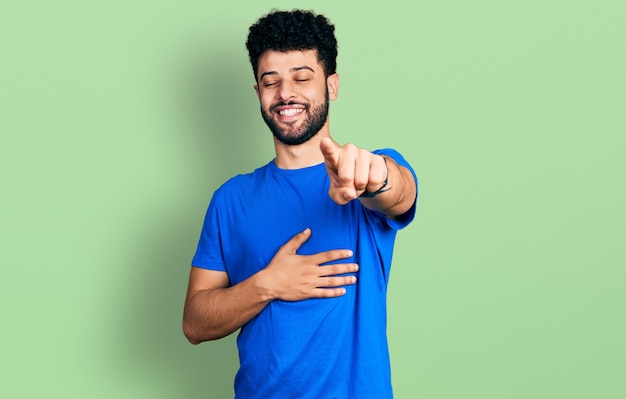 Giovane uomo arabo con la barba che indossa una maglietta blu casual che ride di te puntando il dito verso la telecamera con l'espressione di vergogna del corpo