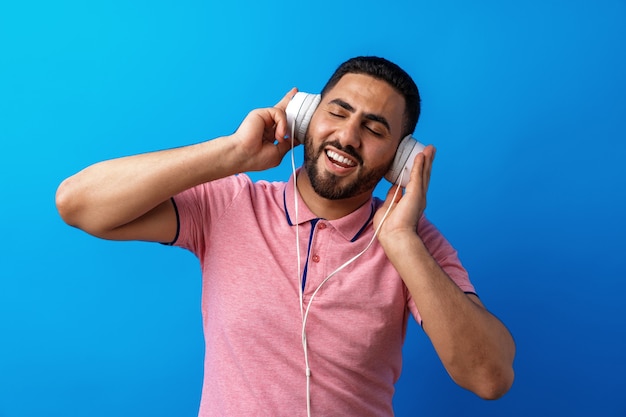 Giovane uomo arabo felice con le cuffie che ascolta la musica contro il fondo blu