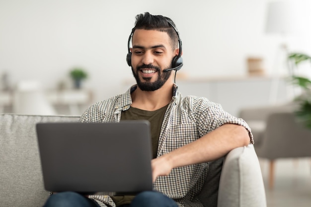 Giovane uomo arabo felice in cuffia utilizzando il pc portatile che lavora online da casa