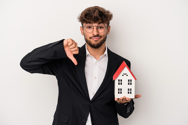Giovane uomo arabo immobiliare che tiene una casa modello isolata su sfondo isolato che mostra un gesto di antipatia, pollice verso. Concetto di disaccordo.