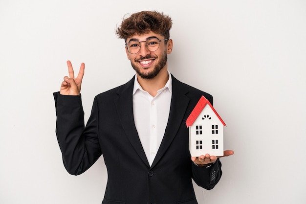 Giovane uomo arabo immobiliare che tiene una casa modello isolata su sfondo isolato gioiosa e spensierata che mostra un simbolo di pace con le dita.