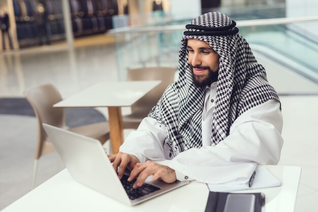 Giovane uomo arabo in sciarpa che utilizza computer portatile nel caffè.