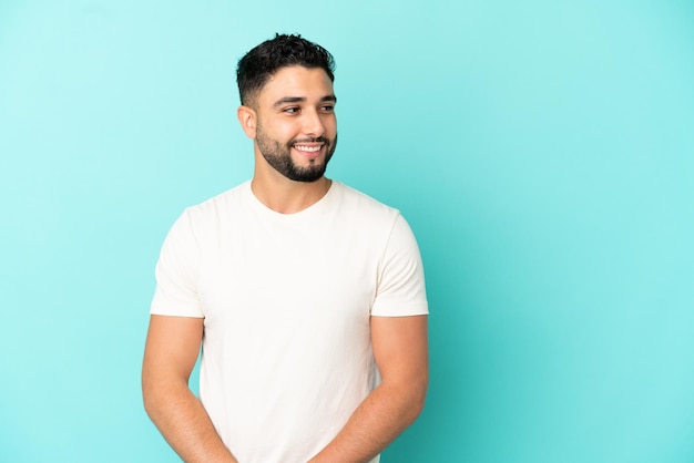 Giovane uomo arabo isolato su sfondo blu guardando di lato e sorridente