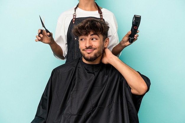 Giovane uomo arabo pronto a tagliarsi i capelli isolato su sfondo blu toccando la nuca, pensando e facendo una scelta.