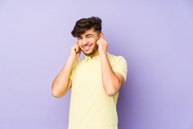 Giovane uomo arabo su un muro viola che copre le orecchie con le mani.