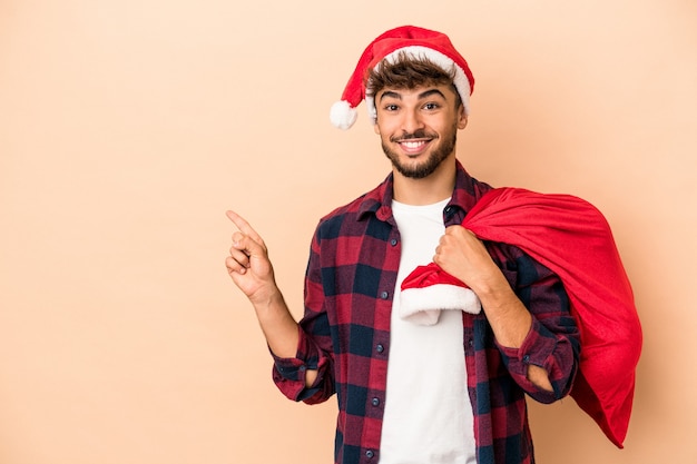 Giovane uomo arabo travestito da babbo natale isolato su sfondo beige sorridente e rivolto da parte, mostrando qualcosa in uno spazio vuoto.