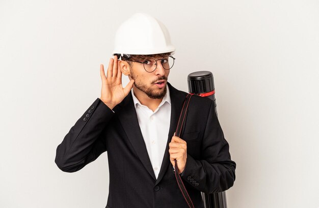 Giovane uomo architetto di razza mista che indossa un casco isolato su sfondo grigio cercando di ascoltare un pettegolezzo.