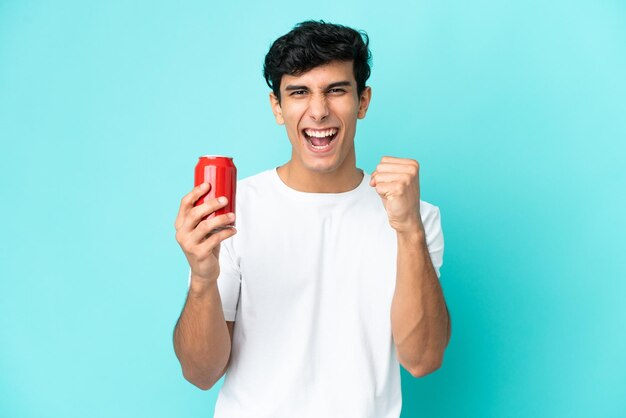 Giovane uomo argentino che tiene un rinfresco isolato su sfondo blu che celebra una vittoria nella posizione del vincitore