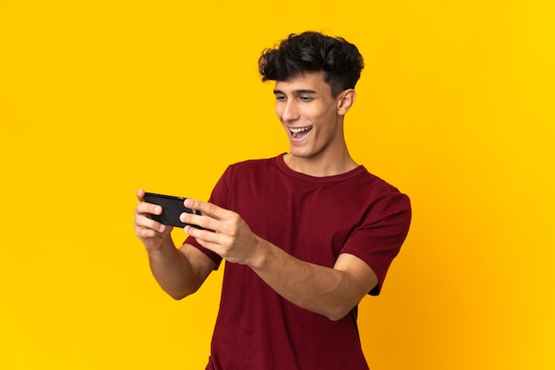 Giovane uomo argentino isolato sulla parete gialla che gioca con il telefono cellulare