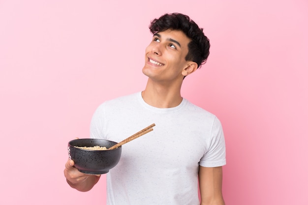 Giovane uomo argentino sopra la parete bianca isolata che cerca mentre sorridendo mentre tenendo una ciotola di tagliatelle con le bacchette