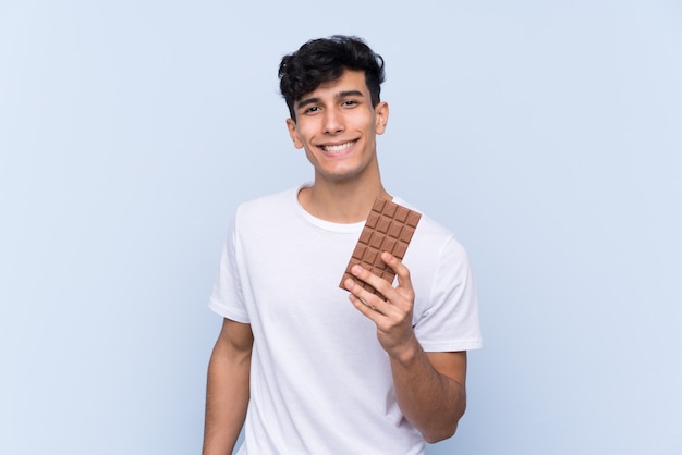 Giovane uomo argentino sopra la parete blu isolata che prende una compressa del cioccolato e felice