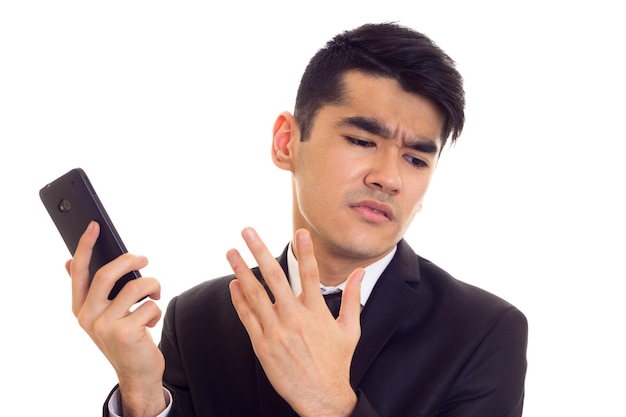 Giovane uomo arrabbiato con capelli neri in camicia bianca e abito nero con cravatta usando il suo telefono