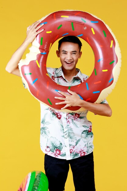 Giovane uomo asiatico attraente che indossa cappello e camicia hawaiana bianca con motivi floreali rossi e verdi e anello di nuotata a ciambella davanti al viso andando in vacanza al mare su sfondo giallo.