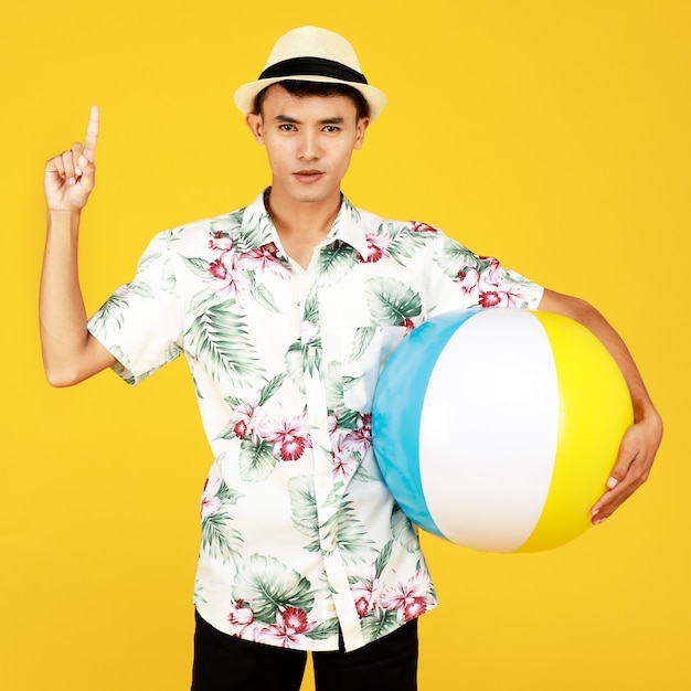 Giovane uomo asiatico attraente in camicia hawaiana bianca che indossa cappello bianco che tiene palla da spiaggia su sfondo giallo. Concetto per le vacanze al mare.