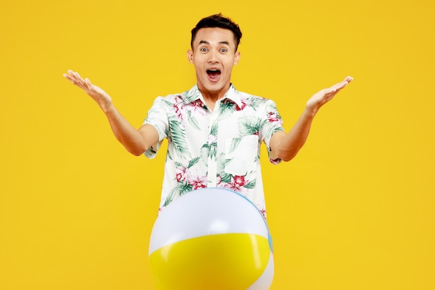 Giovane uomo asiatico attraente in camicia hawaiana bianca che tiene palla da spiaggia su sfondo giallo. Concetto per le vacanze al mare.