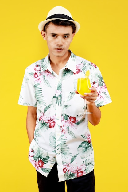 Giovane uomo asiatico attraente in camicia hawaiana bianca che tiene un bicchiere di succo d'arancia su sfondo giallo. Concetto per le vacanze al mare.