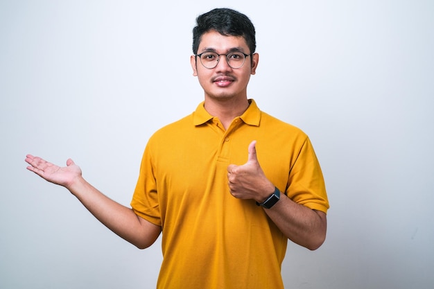 Giovane uomo asiatico bello che indossa una camicia casual che mostra la mano del palmo e che fa gesto giusto con i pollici in su sorridendo felice e allegro su sfondo bianco