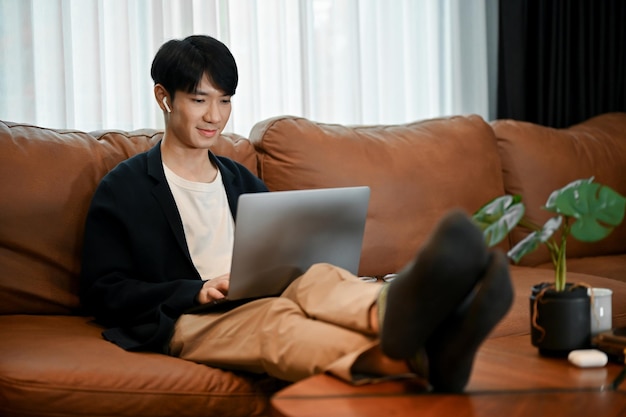 Giovane uomo asiatico bello che si rilassa sul suo divano mentre usa il portatile