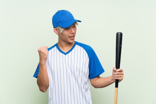 Giovane uomo asiatico che gioca a baseball sopra fondo verde isolato che celebra una vittoria