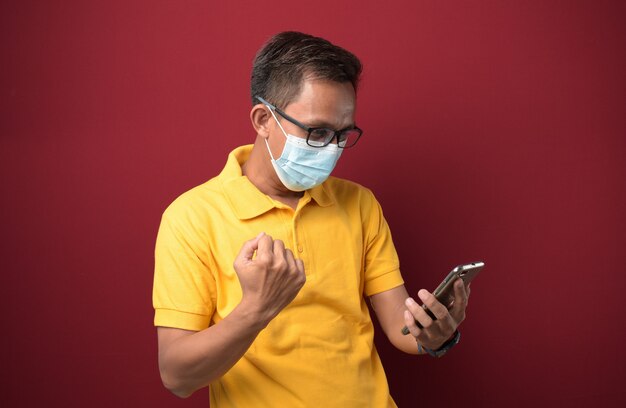 Giovane uomo asiatico che indossa una maschera medica utilizzando lo smartphone che urla orgoglioso e celebra la vittoria.