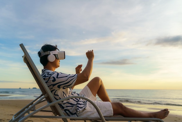 Giovane uomo asiatico che utilizza occhiali per realtà virtuale per riunioni di lavoro sulla spiaggia tropicale su uno sfondo bellissimo mare e cielo