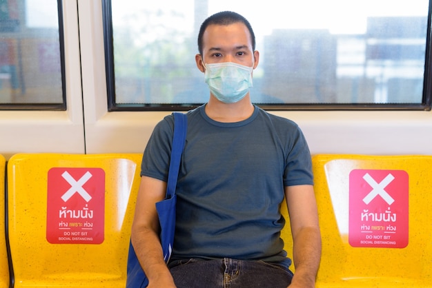 Giovane uomo asiatico con la maschera che si siede con la distanza all'interno del treno