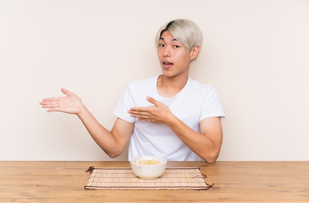 Giovane uomo asiatico con ramen in una tabella che estende le mani al lato per invitare a venire