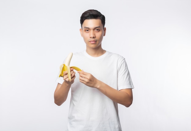 Giovane uomo asiatico in banana bianca della sbucciatura della maglietta e guardare alla macchina fotografica isolata sulla parete bianca