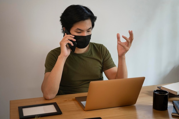 Giovane uomo asiatico in maschera facciale che parla sul cellulare e utilizza il computer portatile al bar