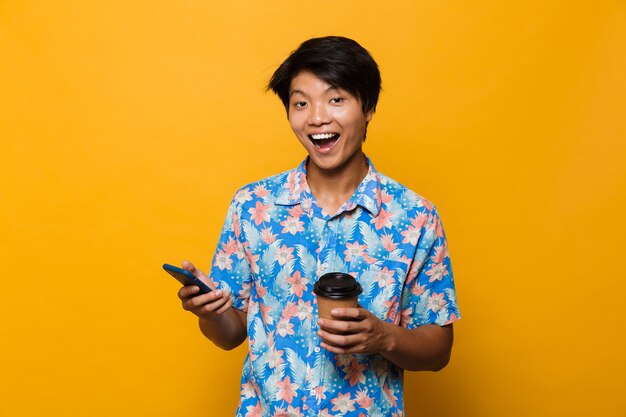 Giovane uomo asiatico in piedi isolato su uno spazio giallo utilizzando il telefono cellulare bere caffè.