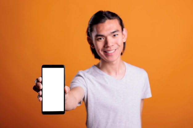 Giovane uomo asiatico sorridente che fa pubblicità allo schermo vuoto dello smartphone, adolescente che tiene il telefono cellulare con il posto per testo. Persona attraente che presenta telefono con vista ravvicinata su display vuoto