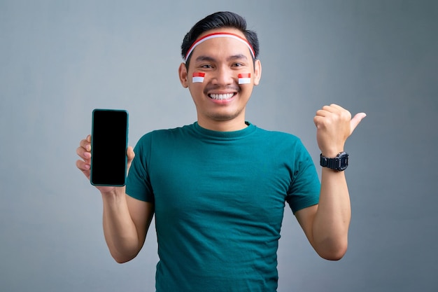 Giovane uomo asiatico sorridente che mostra il telefono cellulare con lo schermo vuoto che consiglia l'app mobile che indica il pollice da parte allo spazio della copia isolato su fondo grigio concetto di celebrazione del giorno dell'indipendenza indonesiana