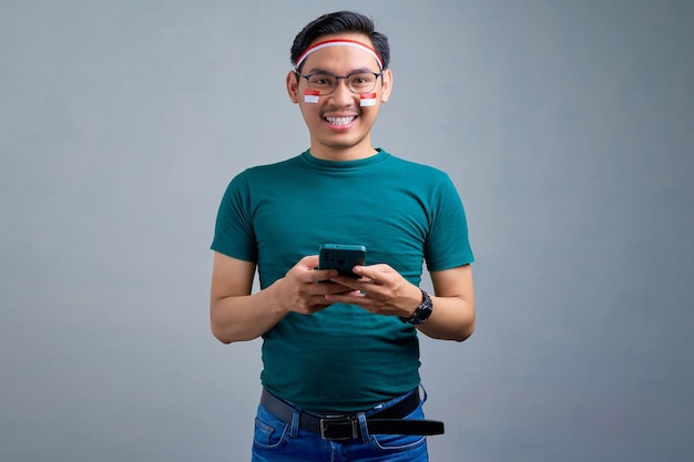 Giovane uomo asiatico sorridente in maglietta casual che tiene il telefono cellulare e guarda la fotocamera isolata su sfondo grigio concetto di celebrazione del giorno dell'indipendenza indonesiana