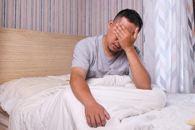 Giovane uomo asiatico stanco seduto a letto a casa ragazzo millenario esausto che si sveglia nella sensazione della camera da letto