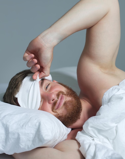 Giovane uomo assonnato bello felice nel letto in camera da letto a casa la mattina sdraiato sotto la coperta bianca godersi il sonno riposante bene nella maschera notturna sugli occhi del viso Foto verticale