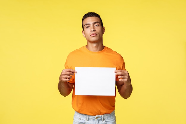 Giovane uomo atletico bello motivato e sicuro di sé serio in maglietta arancione, tenere carta bianca e guardare la macchina fotografica assertiva, promuovere qualcosa di scritto, pubblicizzare il concetto, sfondo giallo