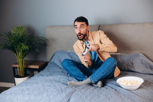 Giovane uomo attraente che gioca a un gioco per console davanti alla telecamera, battendo il suo avversario online. Uomo bianco sul divano con pop corn. Concetto di competizione e-sport.