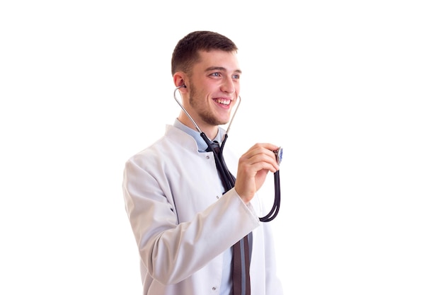 Giovane uomo attraente con capelli castani in cravatta blu e abito da medico utilizzando lo stetoscopio in studio