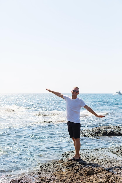 Giovane uomo attraente in occhiali da sole in una maglietta bianca e pantaloncini si trova sulla riva del Mar Mediterraneo