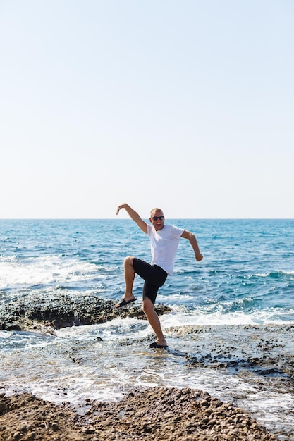 Giovane uomo attraente in occhiali da sole in una maglietta bianca e pantaloncini si trova sulla riva del Mar Mediterraneo