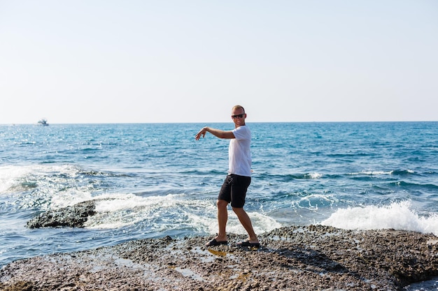 Giovane uomo attraente in occhiali da sole in una maglietta bianca e pantaloncini si trova sulla riva del Mar Mediterraneo