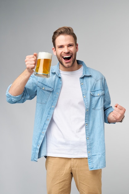 Giovane uomo azienda indossa jeans shirt tenendo un bicchiere di birra in piedi sopra il muro grigio.