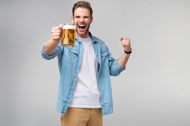 Giovane uomo azienda indossa jeans shirt tenendo un bicchiere di birra in piedi sopra il muro grigio.