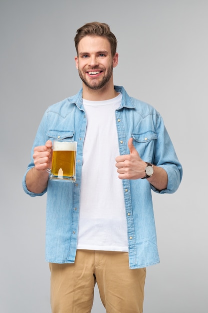 Giovane uomo azienda indossa jeans shirt tenendo un bicchiere di birra in piedi sopra il muro grigio.